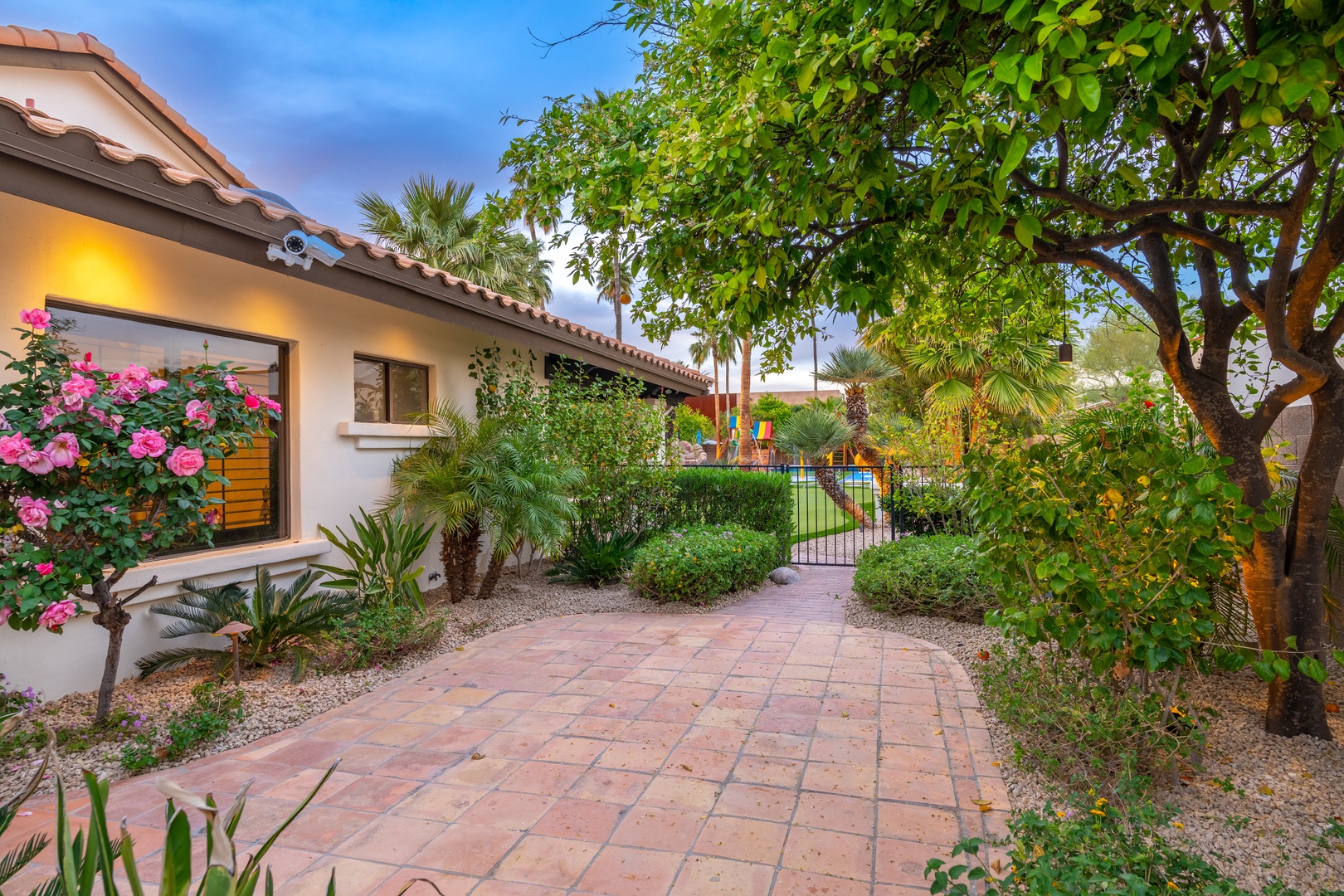 A winding brick pathway leads to the pool through lush landscaping, framed by palm trees and vibrant blooms in the early evening light.