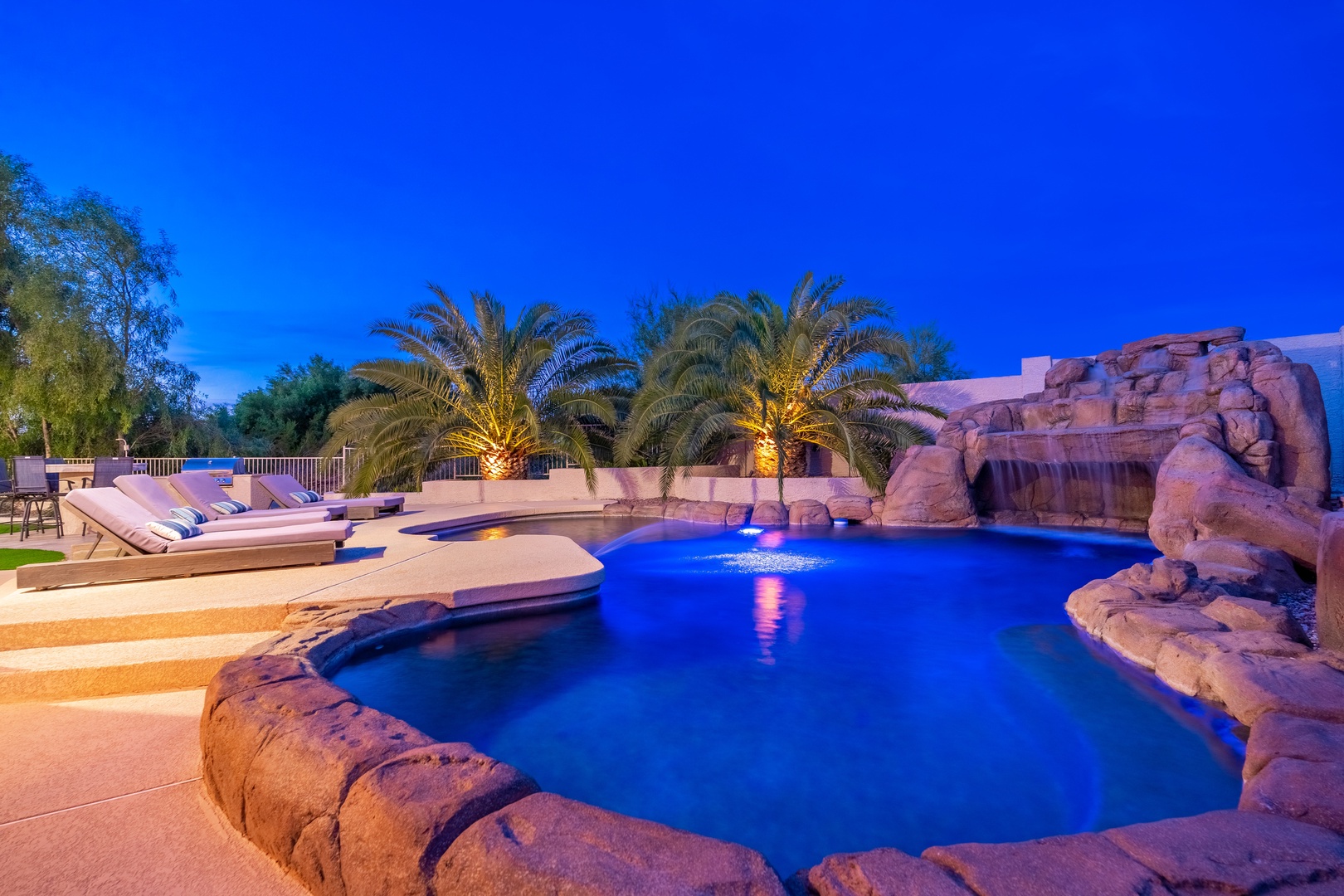 A resort-style pool area featuring a dramatic waterfall grotto, illuminated palms, and loungers—captured at twilight for a stunning backdrop to your desert escape.