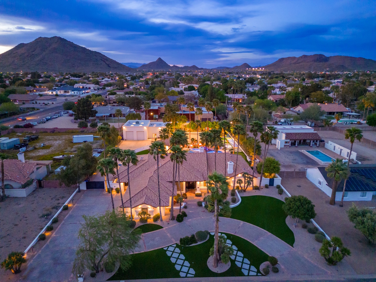 Aerial view of a stunning desert property at dusk, showcasing the villa's elegant architecture, lush palm trees, manicured lawns, and neighboring pool area, all set against dramatic mountain ranges.