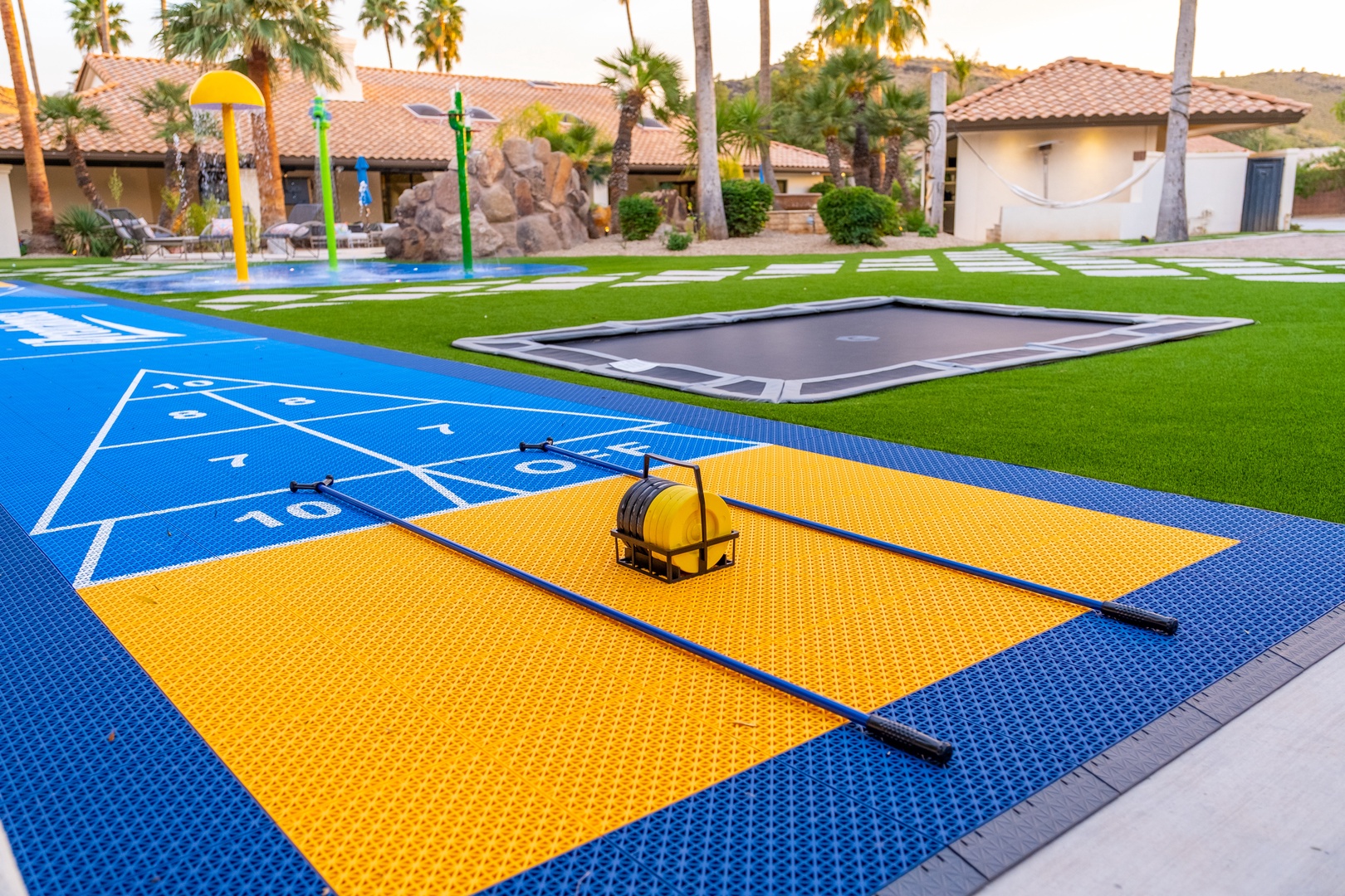 A vibrant splash pad with water features sits alongside a trampoline and creative play zones under sunny skies and swaying palms, perfect for family fun.