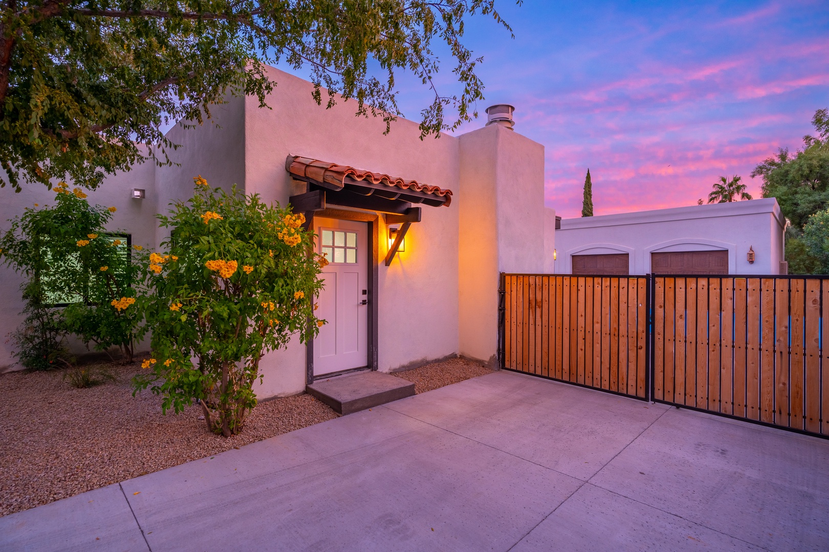 Adobe-style architecture with terra cotta accents and flowering garden greets visitors at the entrance, set against a stunning desert sunset sky.