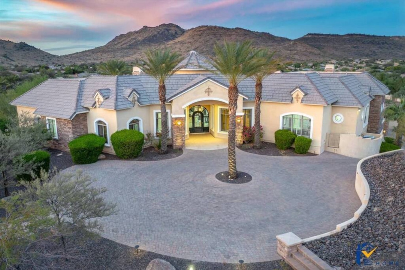 An elegant desert estate nestled against dramatic mountain landscapes, featuring Mediterranean architecture, palm-lined entrance, and spacious paved courtyard at twilight.