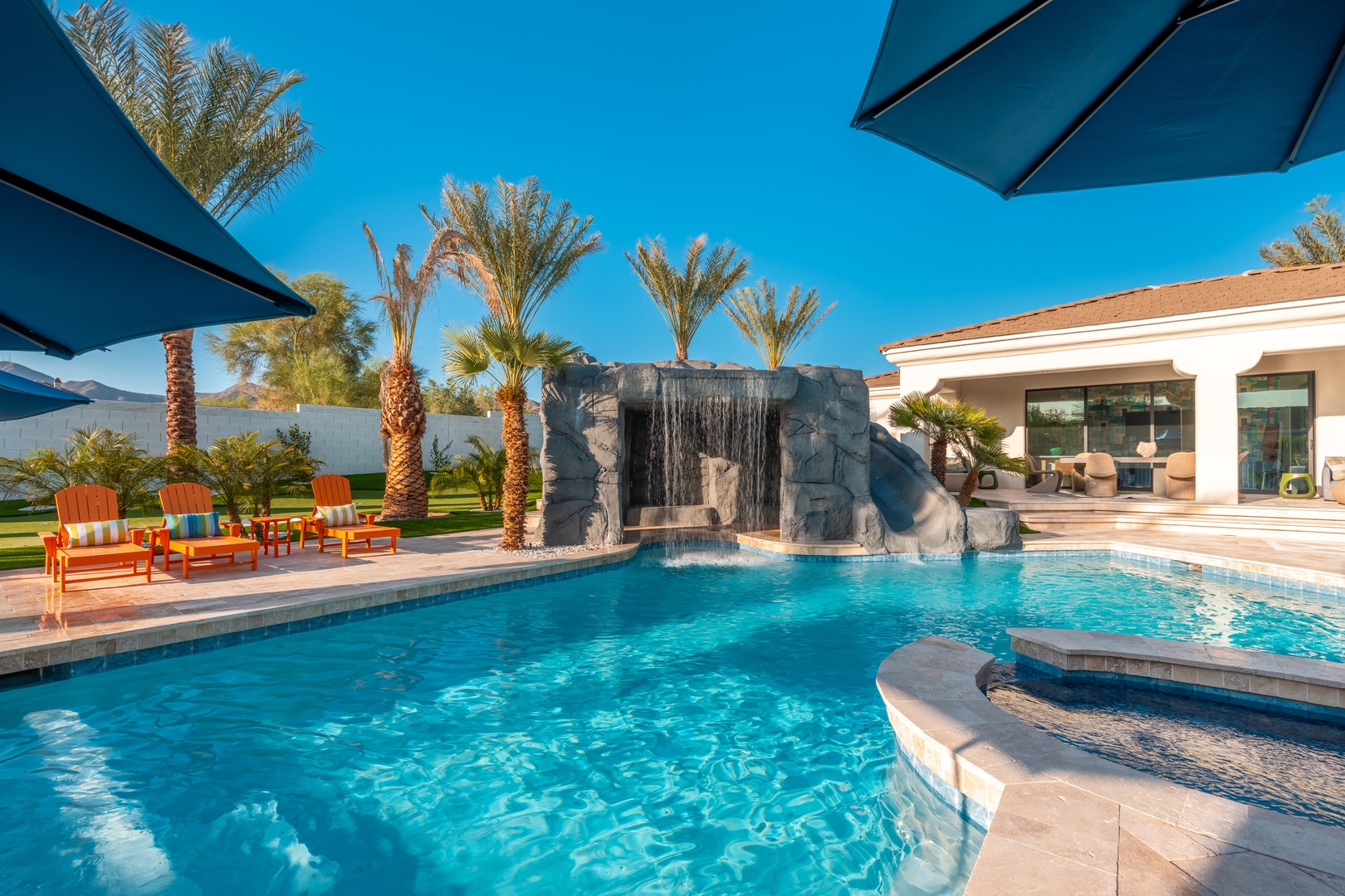 Resort-style pool features a dramatic waterfall grotto, surrounded by swaying palms and mountains—creating a private desert oasis in your own backyard.