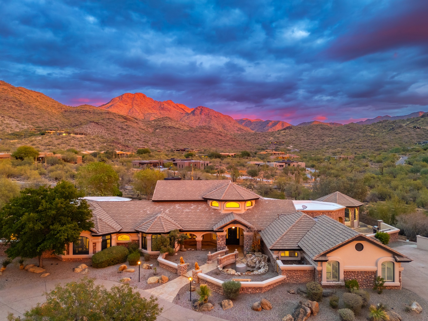 A stunning desert property nestled in the foothills, with dramatic mountain peaks glowing in sunset light beyond the surrounding desert landscape and neighboring homes.