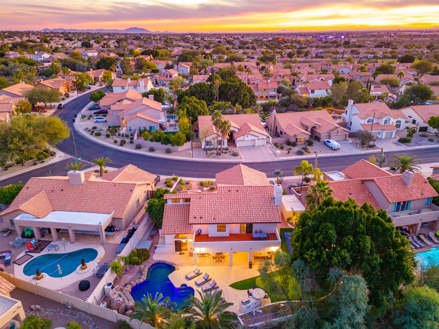 An aerial view captures the property at sunset, situated in a peaceful residential neighborhood with distant mountain vistas and open desert landscape surrounding the community.