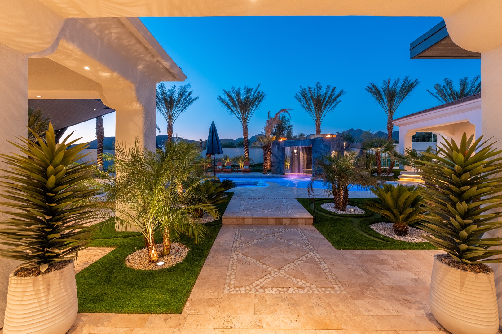 This stunning resort-style property features lush tropical landscaping, elegant architecture, and a sparkling pool area set beneath towering palms, creating an inviting oasis for relaxation and entertainment.