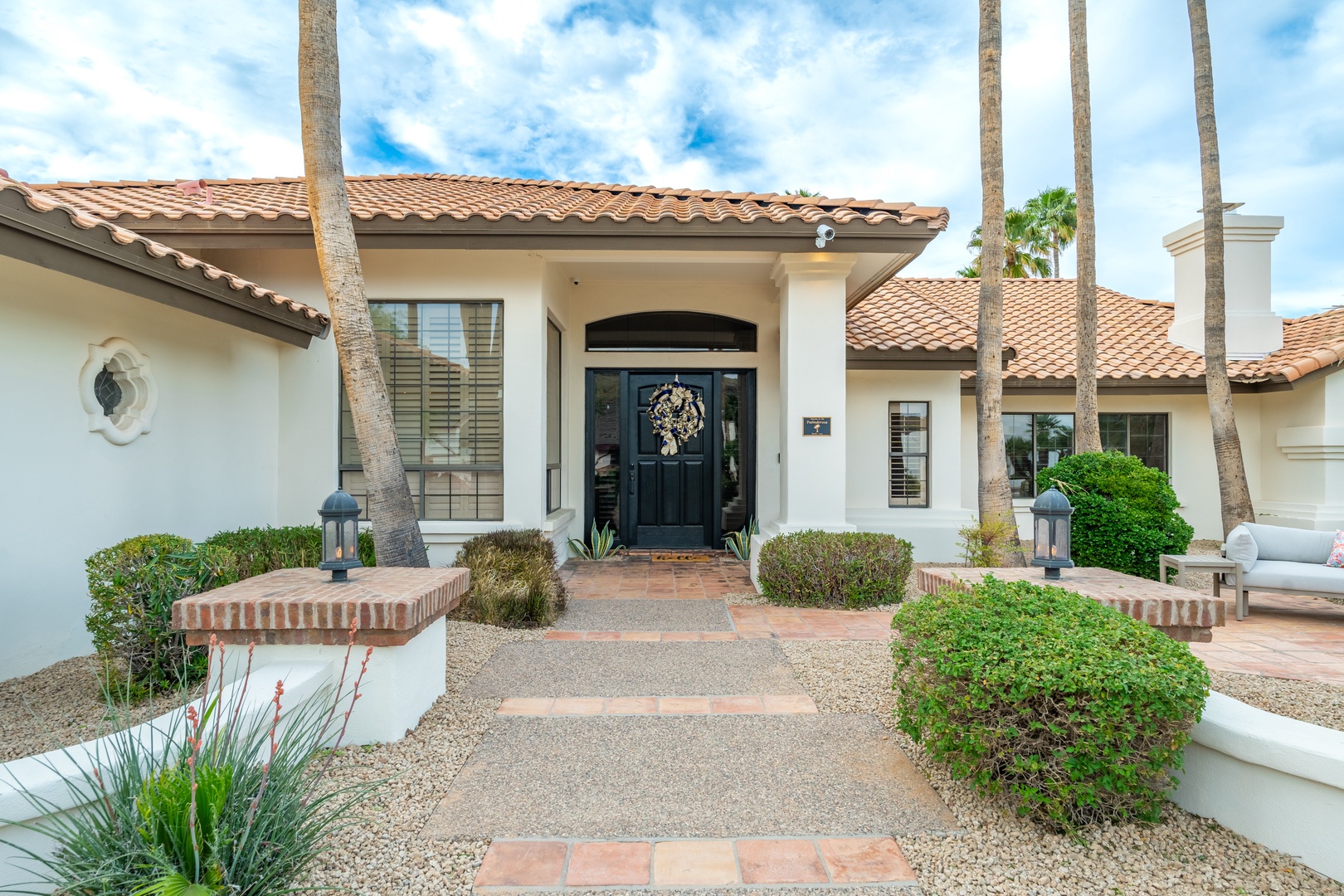 Welcome to your desert oasis. This elegant Mediterranean-style home features a grand arched entry with classic black doors, framed by towering palms and mature desert landscaping that creates a perfect first impression.