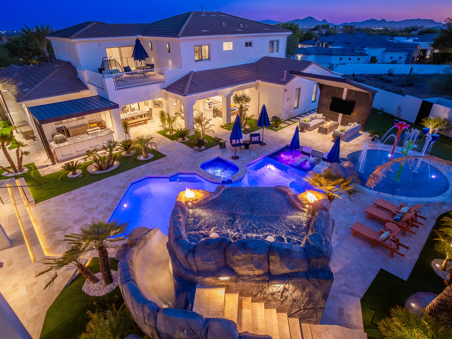 Stunning aerial view of a luxury vacation rental featuring an expansive pool complex with illuminated waterfalls, multiple lounging areas, and tropical landscaping set against mountain vistas at dusk.