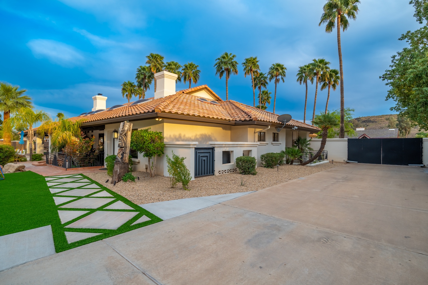 This charming desert-style retreat features a spacious driveway and landscaped entrance, set against a backdrop of swaying palms and mountain views.