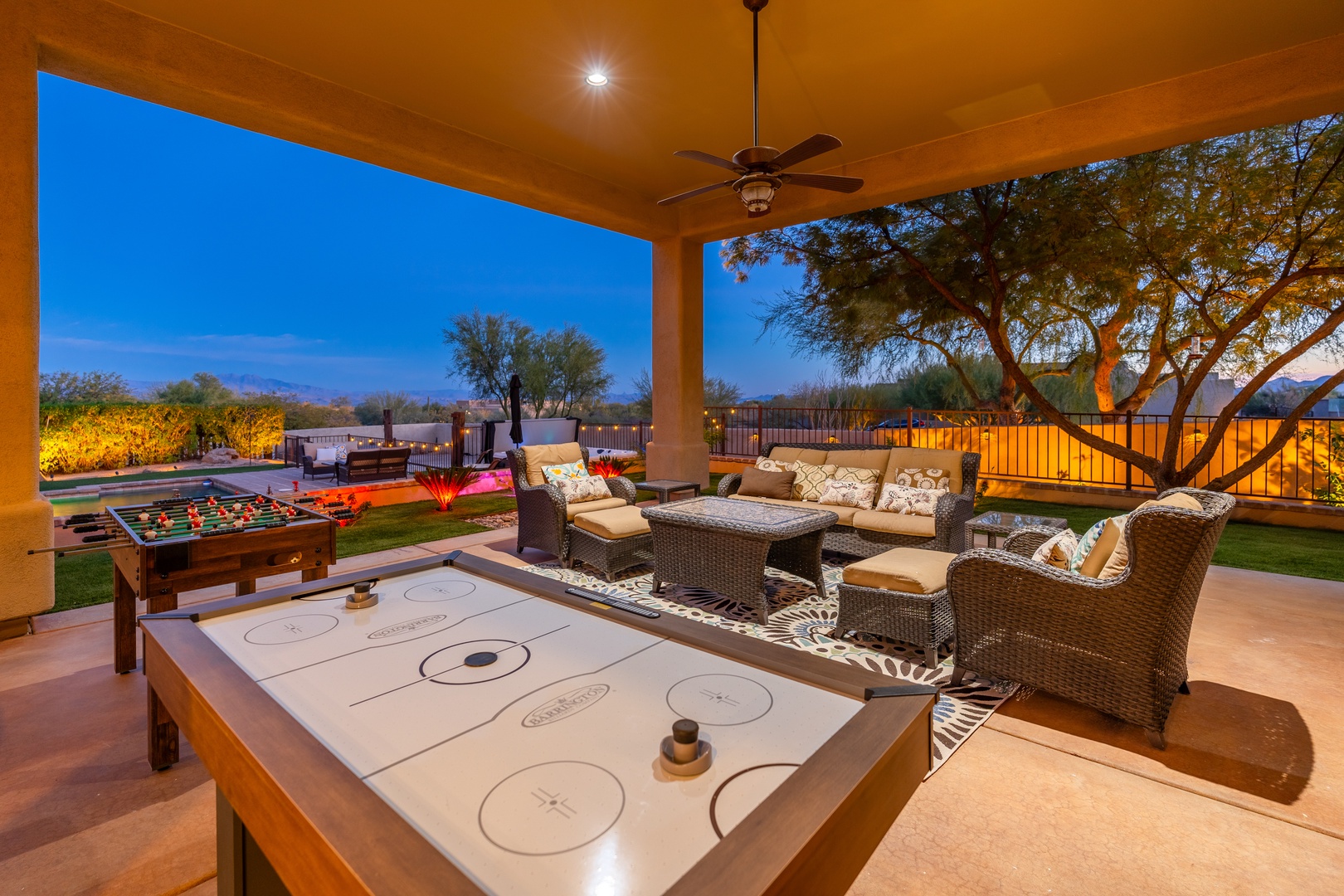Step into your covered patio game zone at dusk, where foosball and air hockey await beneath a glowing sky. Plush wicker seating invites you to unwind as palm trees frame distant mountain views.