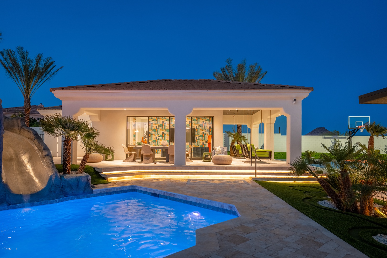 Stunning vacation villa featuring a sparkling private pool, palm-studded gardens, and basketball court set against a twilight sky—a serene desert retreat.