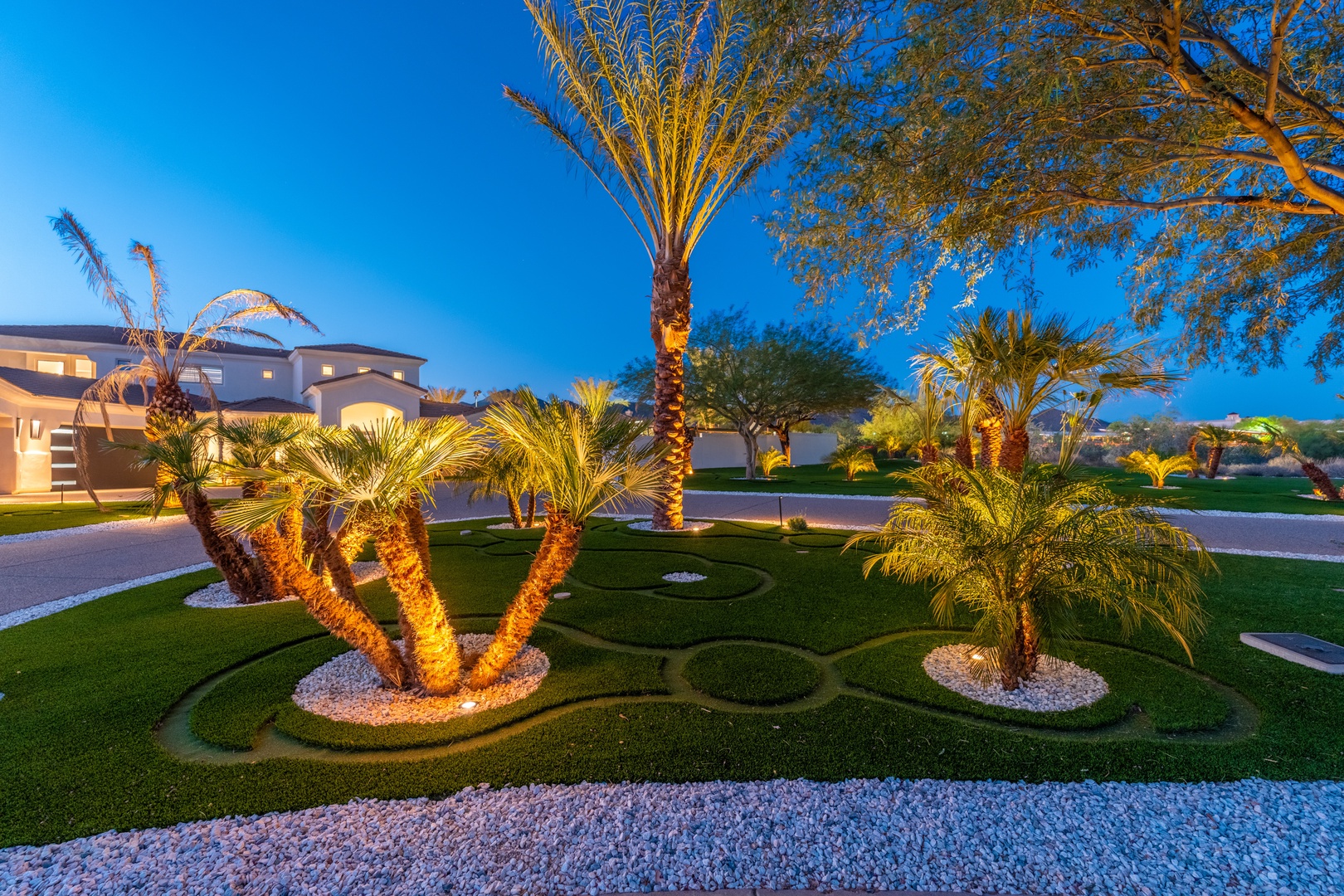 Beautifully landscaped front yard featuring sculptural palm trees, decorative lighting, and manicured lawns at dusk. The property's entrance showcases desert-inspired design in a peaceful residential setting.