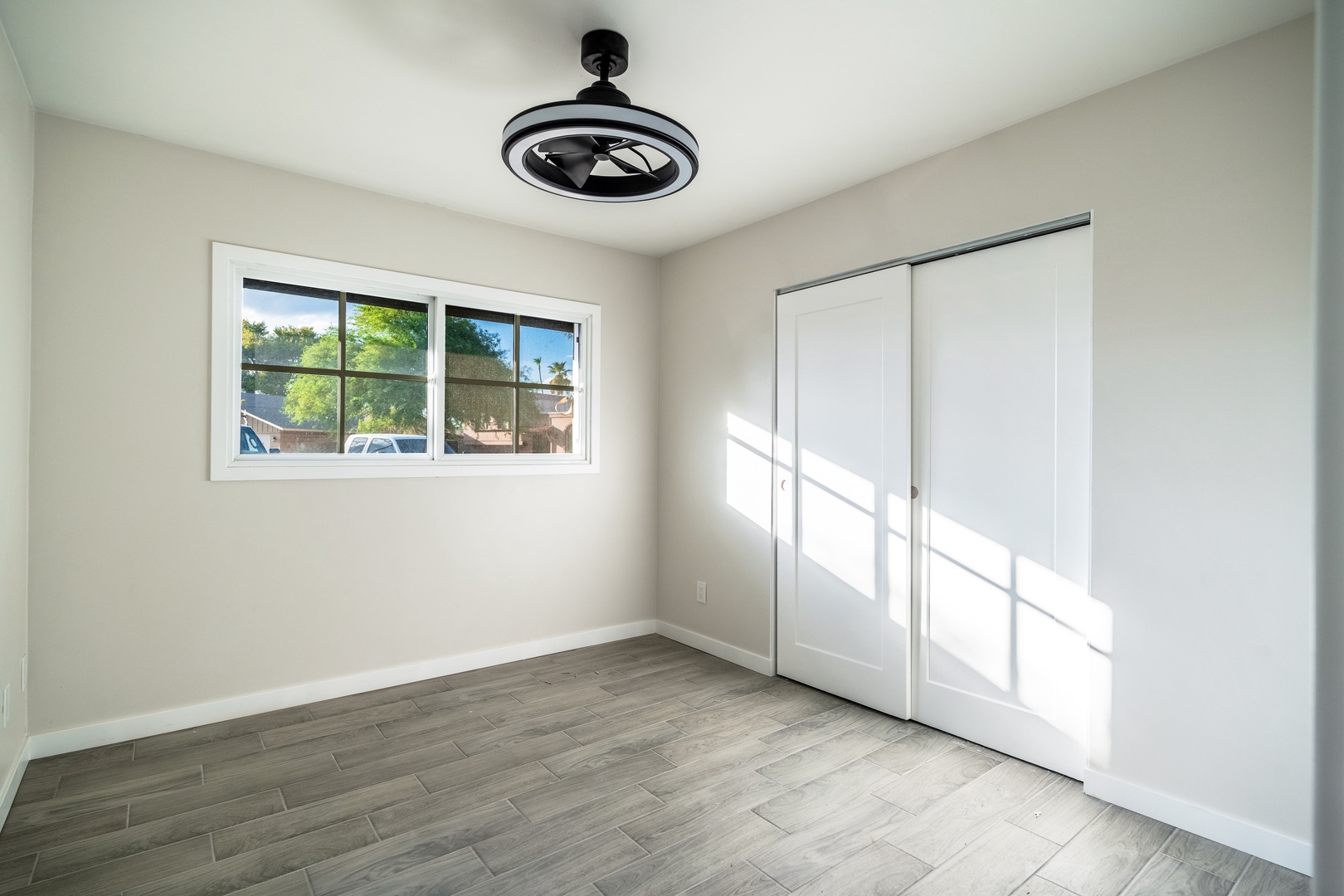An airy, light-filled bedroom with clean lines and modern finishes awaits you. Sunlight streams through the window, highlighting sleek wood-style flooring and a ceiling fan overhead. Your spacious closet keeps everything organized.