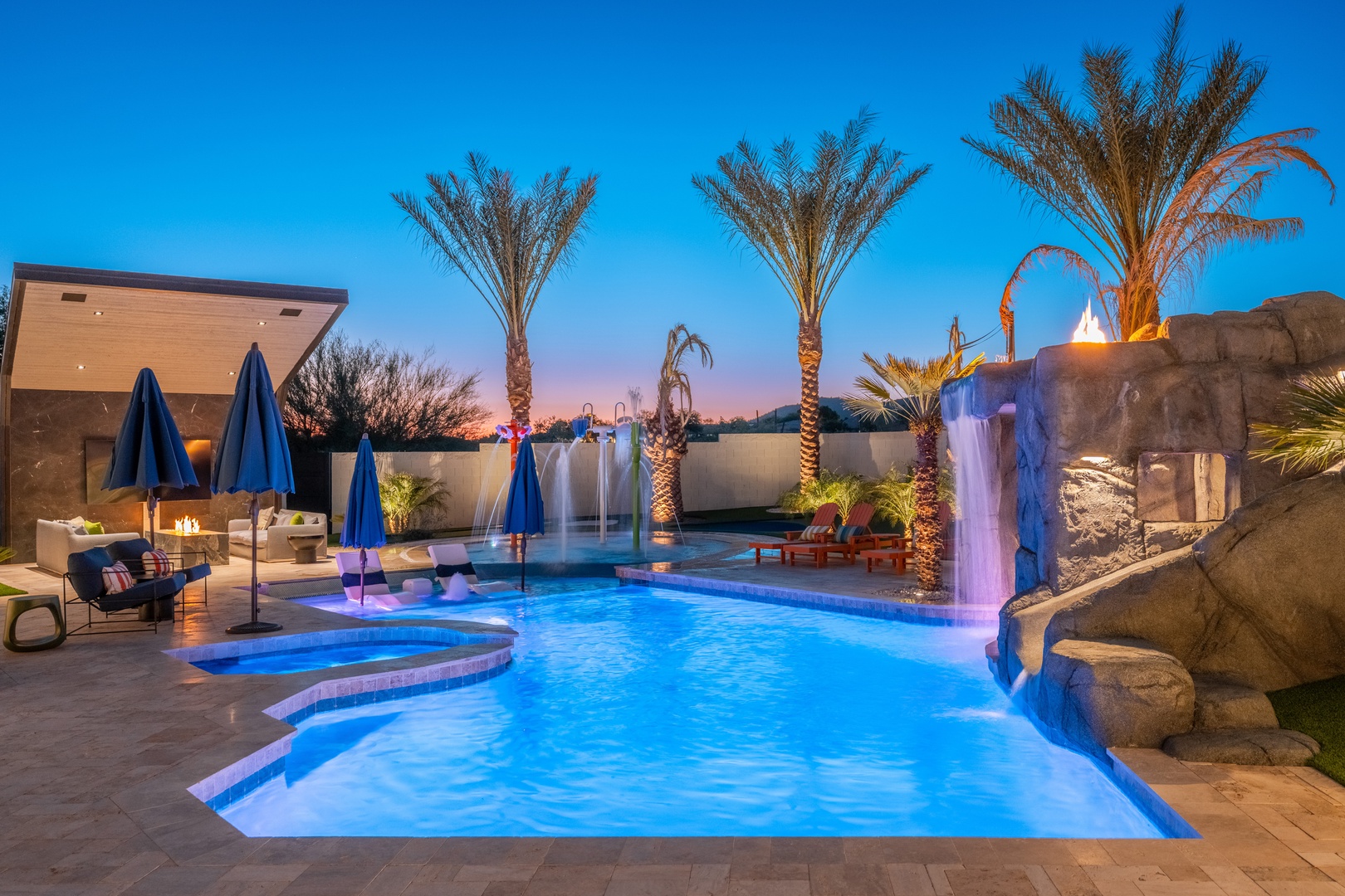 The pool glows under twilight skies, with a dramatic waterfall, cozy fire features, and swaying palms creating a resort-style oasis for your evenings.