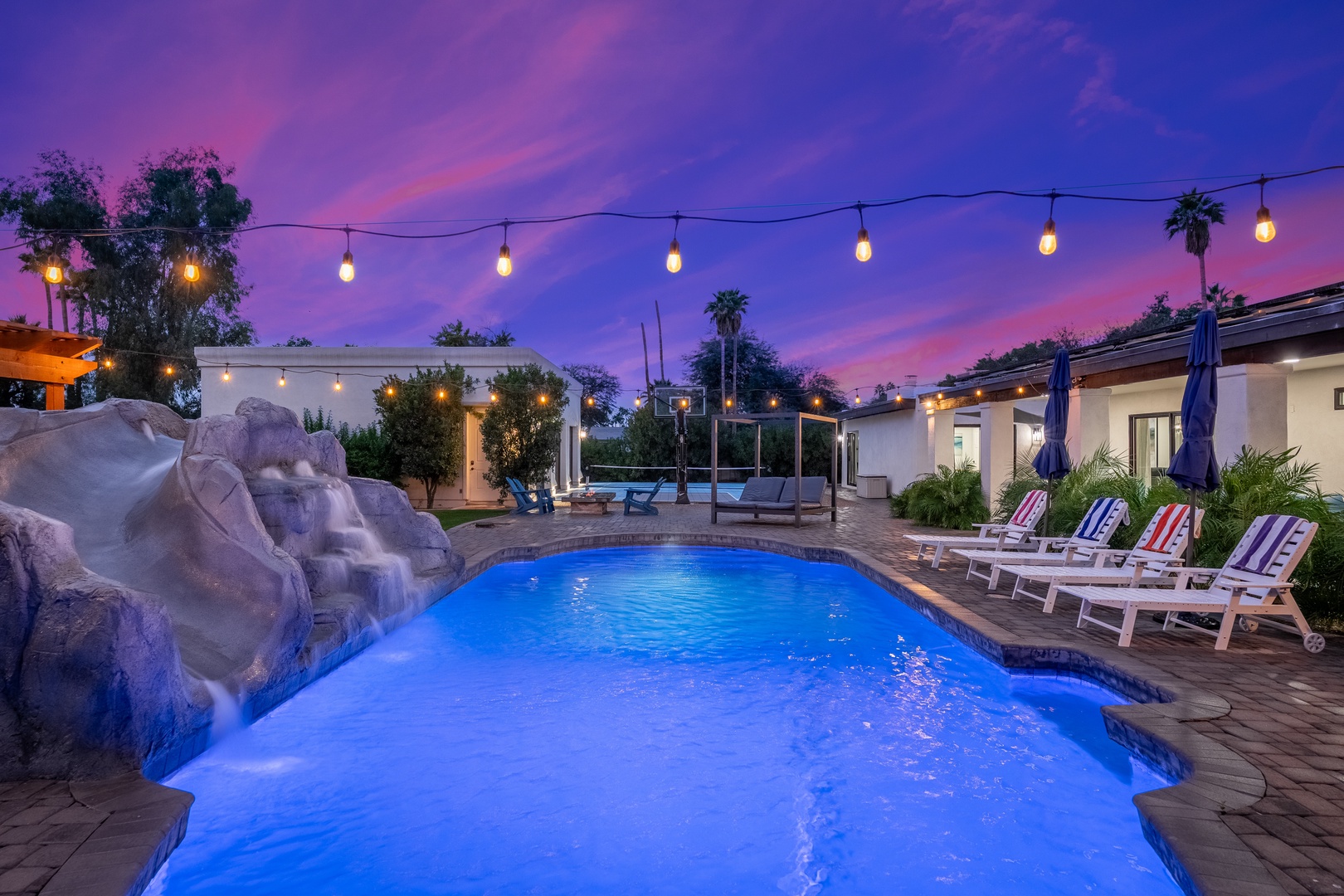 A stunning twilight scene frames the illuminated pool, where colorful lounge chairs await your evening relaxation beneath the glowing string lights.