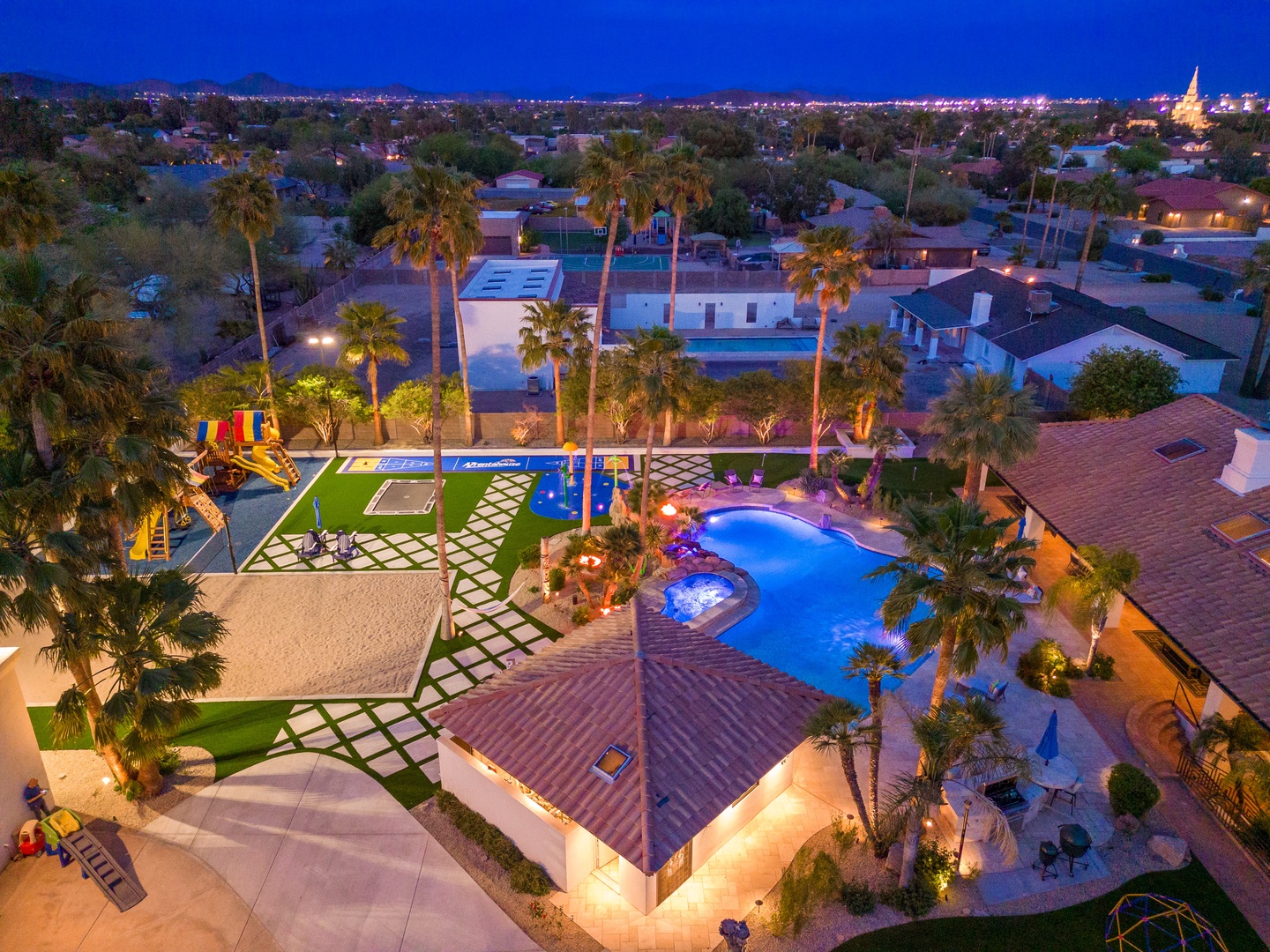 An aerial view showcasing the property's extensive outdoor amenities, including a freeform swimming pool, hot tub, children's playground, and manicured grounds illuminated against the desert evening sky.