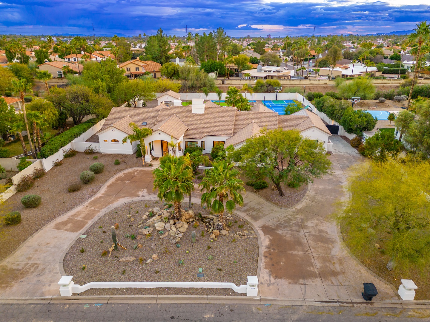 Aerial view of the property showcasing the single-level residence with desert landscaping, palm trees, and surrounding residential neighborhood with nearby amenities.
