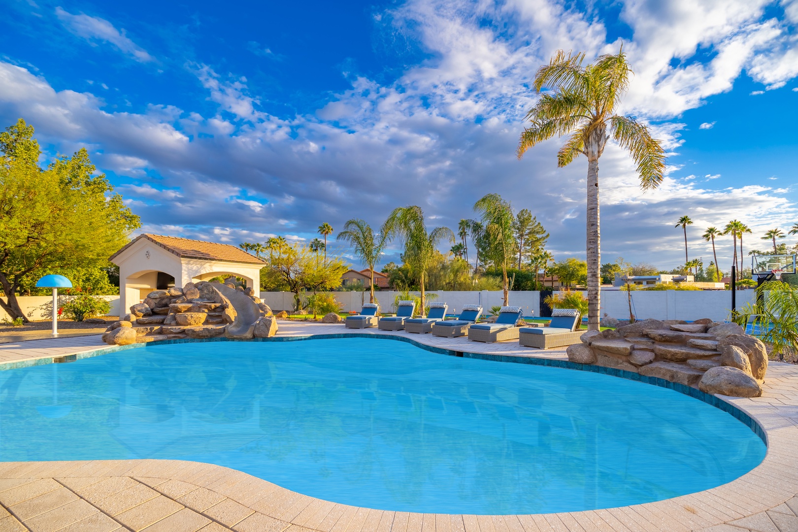 A stunning resort-style pool awaits beneath swaying palms and brilliant blue skies, with lounge chairs and a charming cabana nearby for your relaxation.