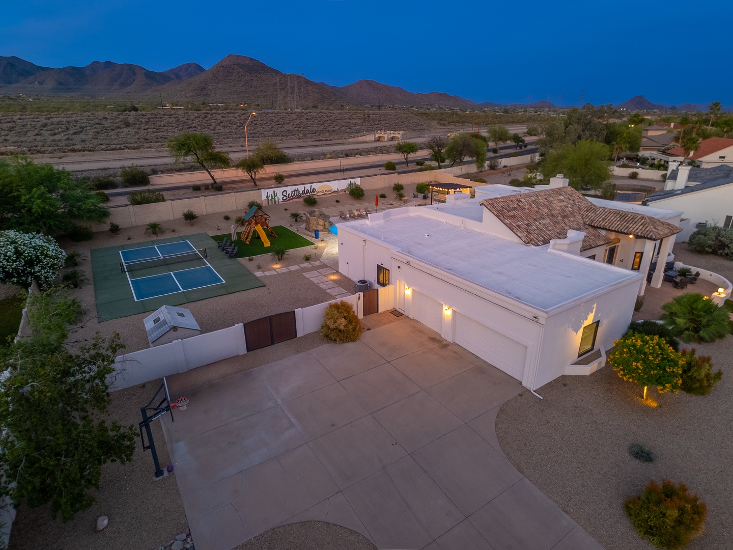 Aerial view of a desert vacation home in Scottsdale, featuring a tennis court, playground, and mountain backdrop at dusk.