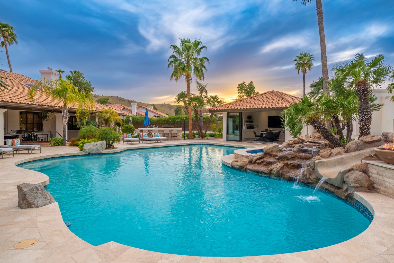 Resort-style pool oasis with tropical palms and cascading rock waterfall, perfect for unwinding under breathtaking desert sunsets.