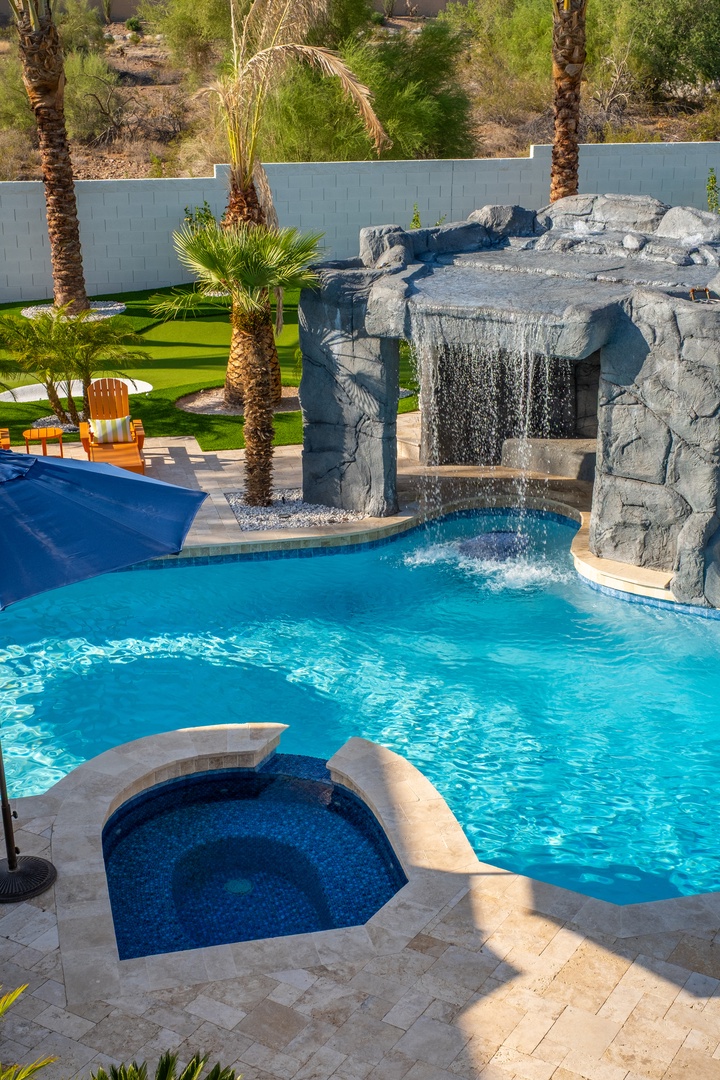 An inviting oasis featuring a sparkling pool with a dramatic stone waterfall, sunken hot tub, and lush landscaping shaded by towering palms—perfect for your desert escape.
