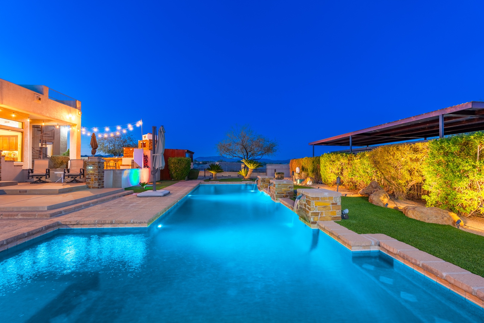 A stunning twilight view of the property's outdoor area, featuring a beautifully lit pool, covered patio with string lights, and lush landscaping creating a perfect evening retreat atmosphere.