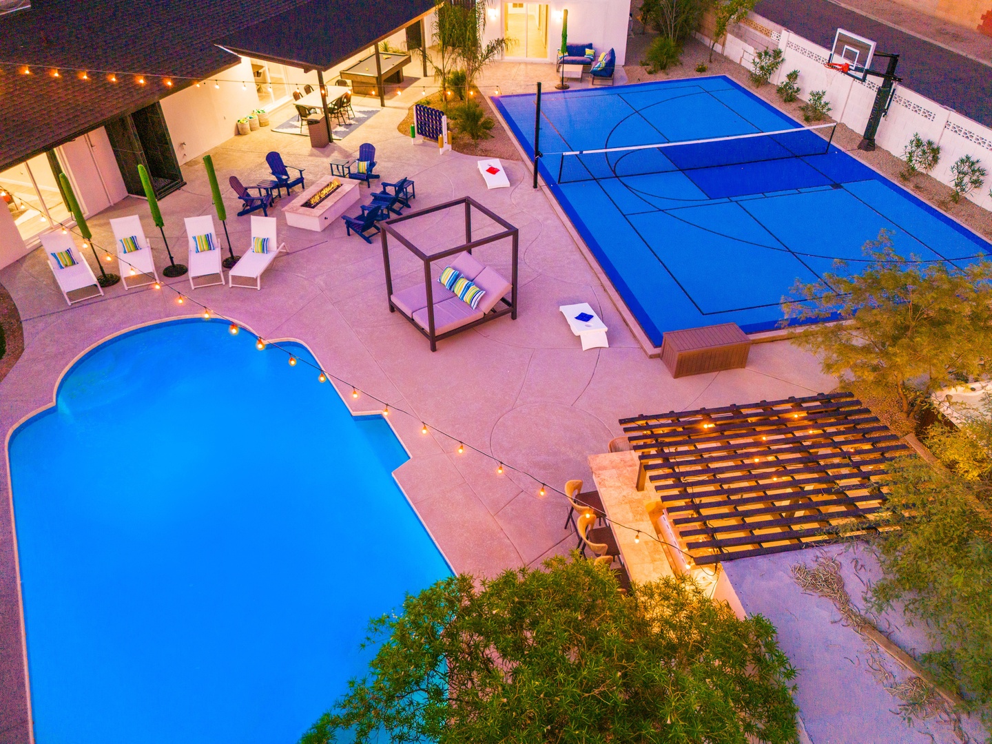 An aerial view captures the property's backyard oasis at twilight, featuring a pristine pool, covered sport court with basketball hoop, lounge seating, and ambient string lighting creating an inviting recreational area.