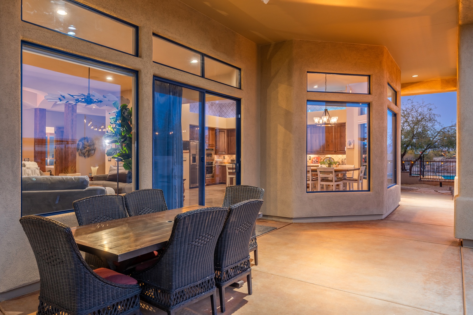Modern southwestern-style retreat with covered patio seating perfect for dining under desert skies. Large windows reveal warm interiors and seamless indoor-outdoor living.