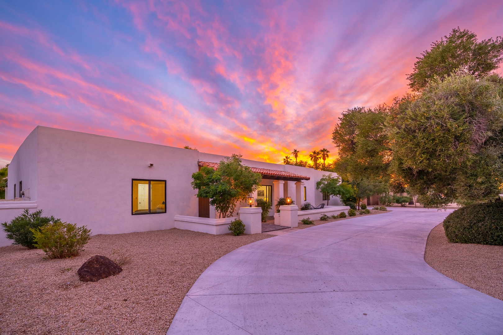 A stunning desert sunset paints the sky in vibrant hues of pink, orange, and purple above this modern southwestern property, surrounded by native landscaping and palm trees.