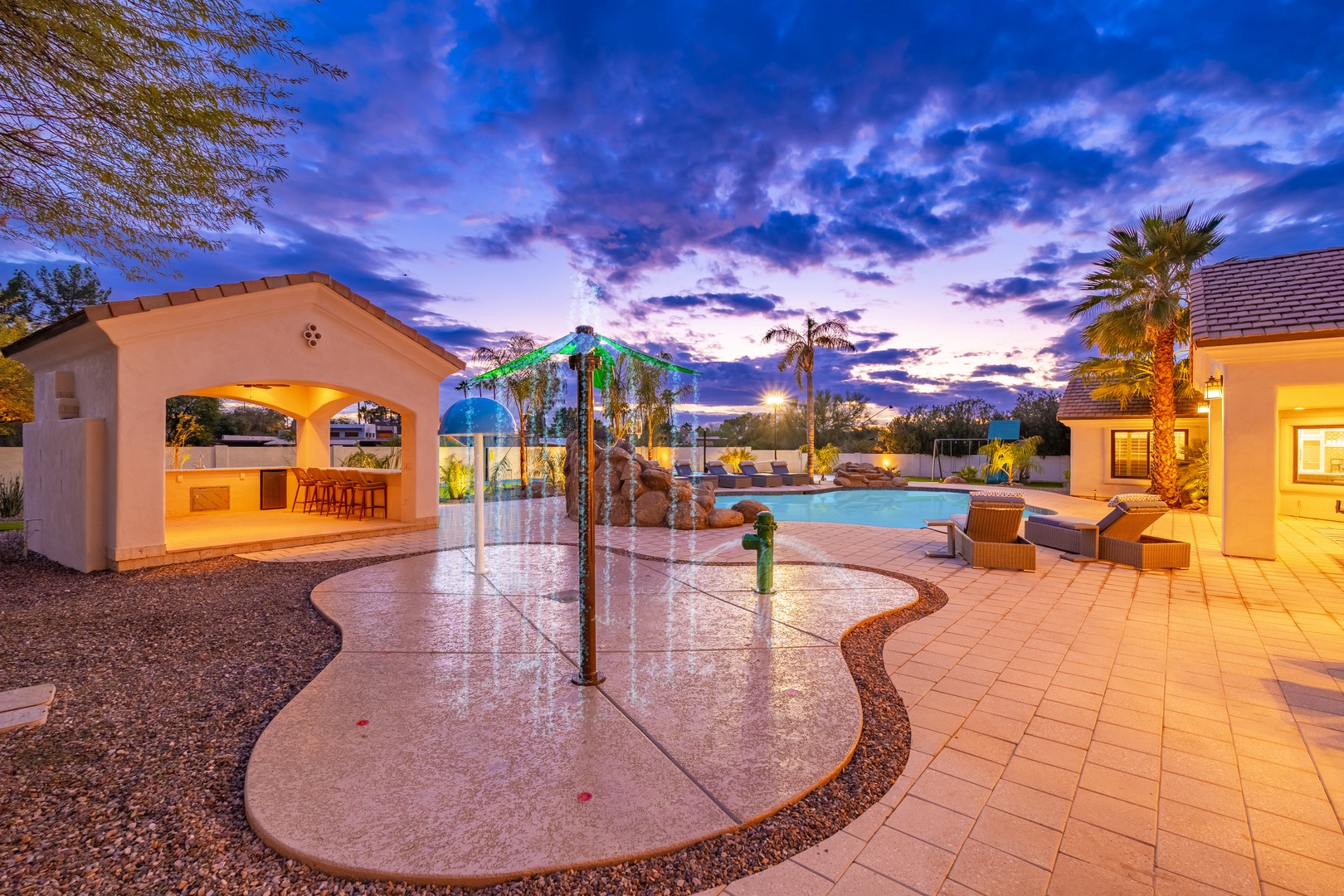 The property features a tropical splash pad, poolside cabana with bar seating, and spacious desert patio—all bathed in stunning twilight hues over palm-studded grounds.