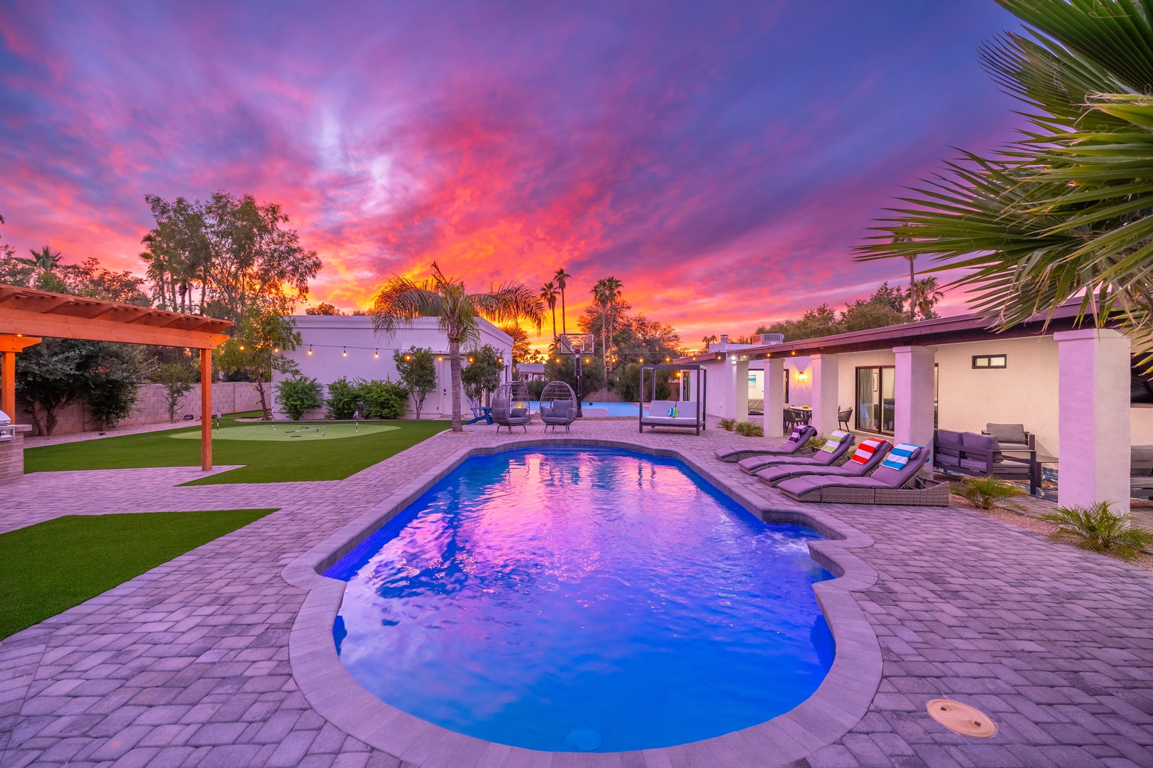 A stunning desert oasis featuring a sparkling pool surrounded by lush palms and manicured lawns, set against a dramatic pink and purple sunset sky.