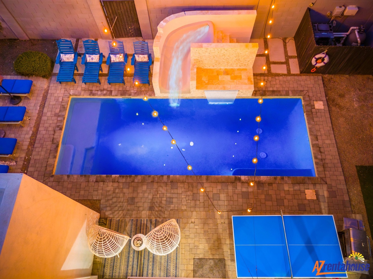 Stunning aerial view showcases the sparkling swimming pool and hot tub area illuminated by warm evening lights, surrounded by blue loungers and entertainment zones.