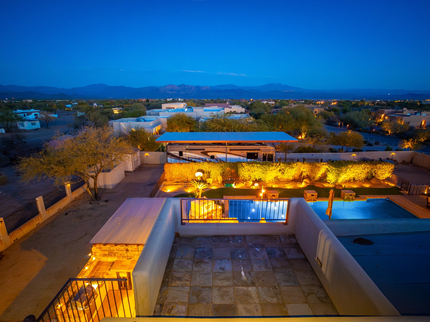 Elevated view of the property at twilight showcasing the illuminated pool, outdoor entertaining spaces, and lush landscaping with mountain and neighborhood views beyond.