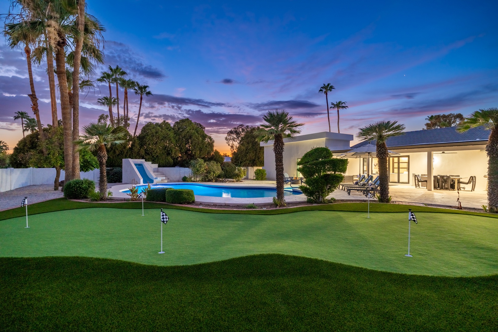 A private putting green, sparkling pool with slide, and modern architecture framed by palms under a stunning desert twilight—an exceptional outdoor retreat awaits.