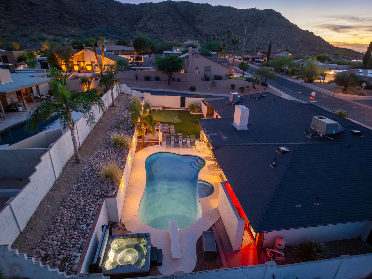 Aerial view captures this vacation retreat at twilight, with mountain backdrop, private pool, hot tub, and spacious yard lit warmly against the desert neighborhood.