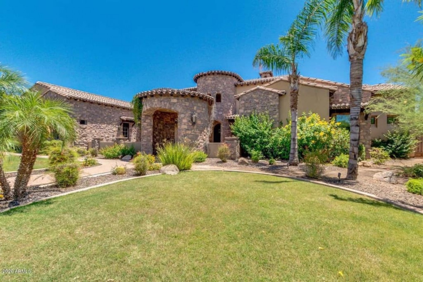 Stunning Mediterranean-style villa with stone facade, clay tile roof, and manicured landscaping featuring palm trees and desert plants.