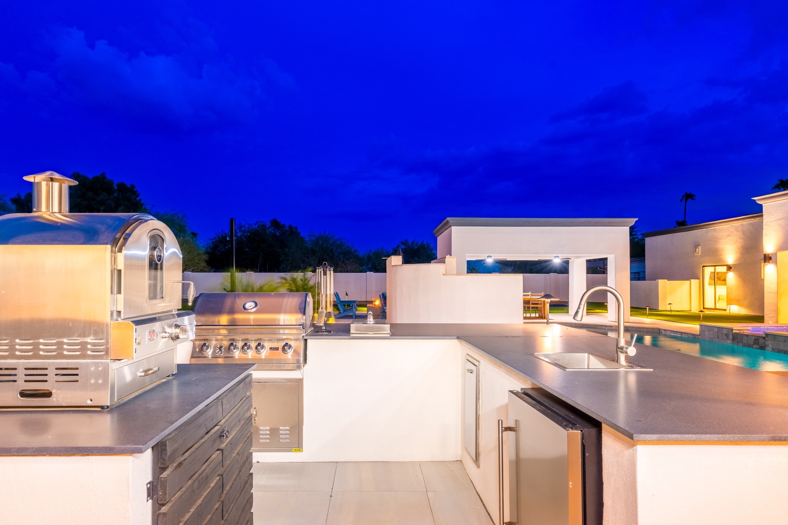 Modern outdoor kitchen with premium grill and sleek countertops, perfect for entertaining under the evening sky while enjoying the illuminated pool and spacious patio beyond.