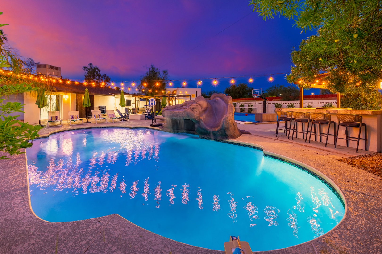 A stunning twilight scene where glowing pool waters mirror vibrant skies, string lights twinkle overhead, and an outdoor bar awaits evening relaxation.