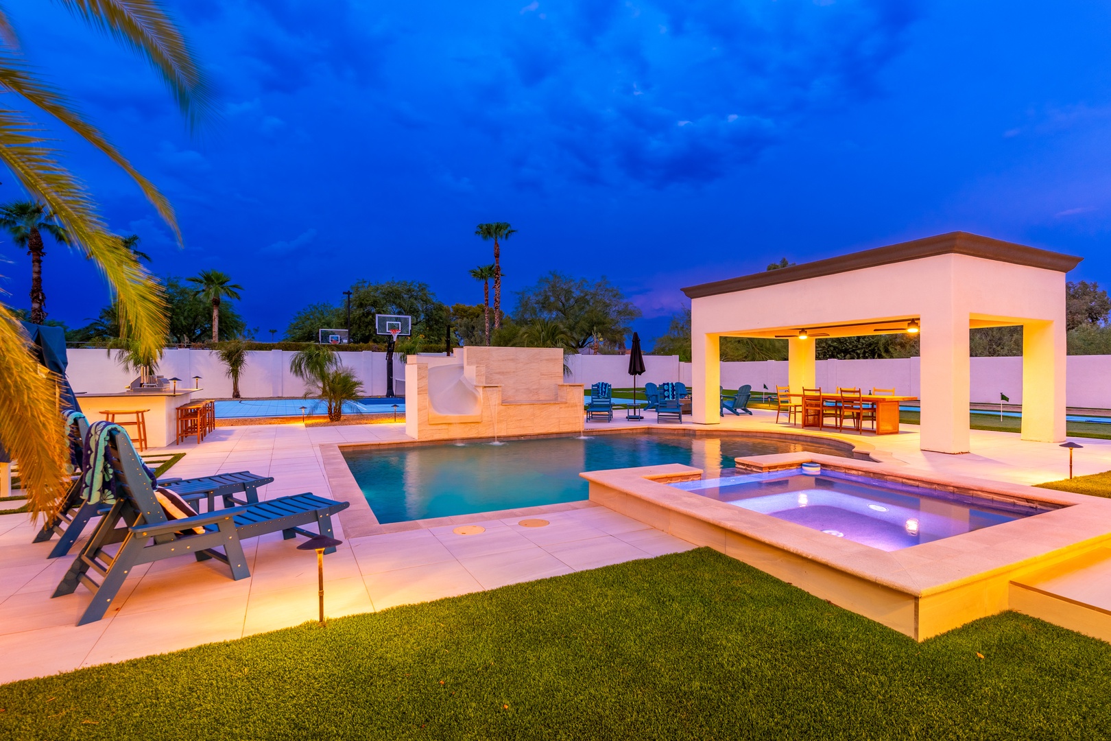 A stunning resort-style backyard captured at twilight, featuring a sparkling pool, elevated spa, covered pavilion, basketball court, and tropical landscaping with palm trees under a vibrant evening sky.