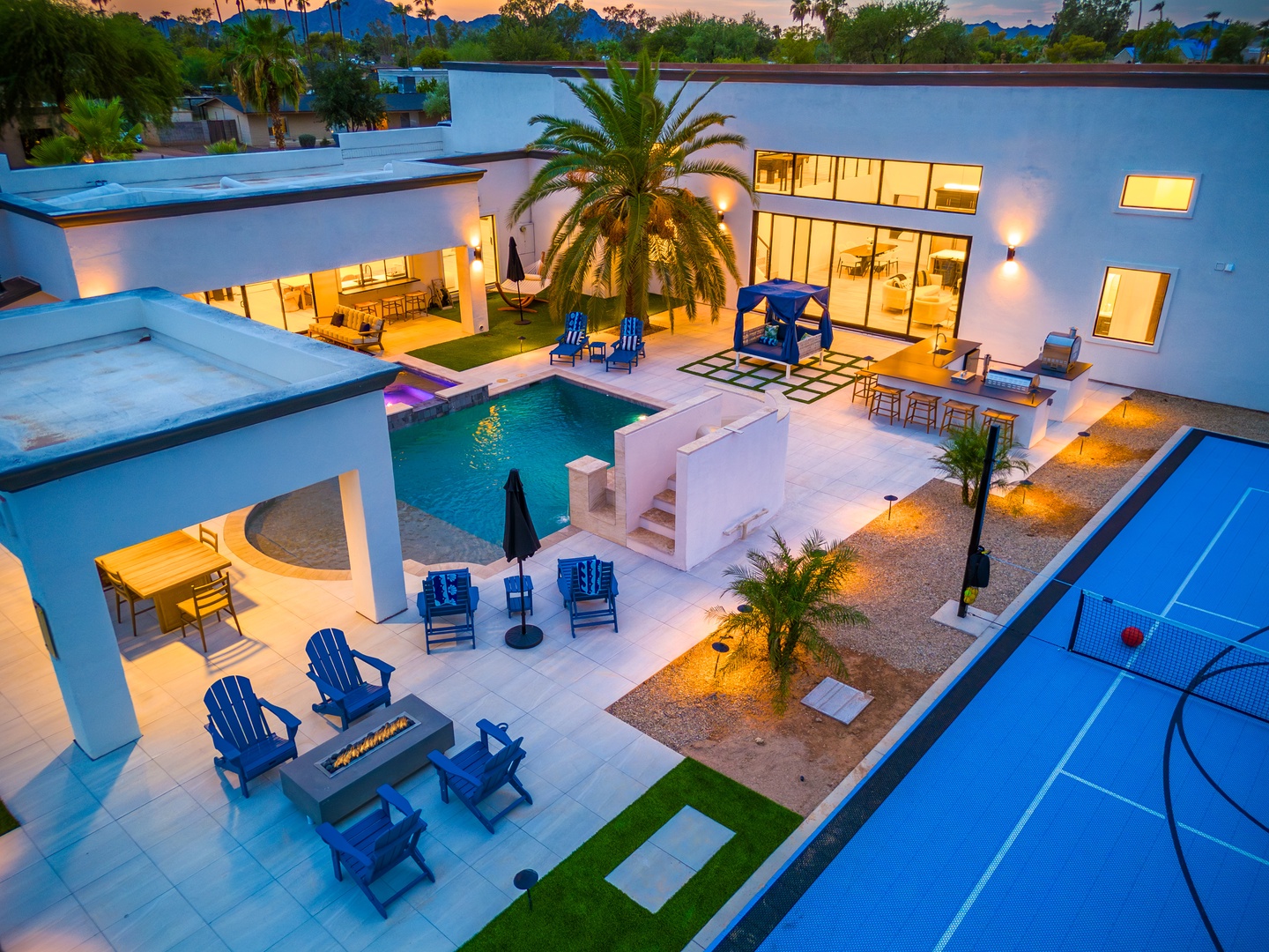 A stunning modern desert oasis awaits, with a sparkling pool, covered patio, sport court, and palm-accented landscaping set against dramatic mountain views at dusk.