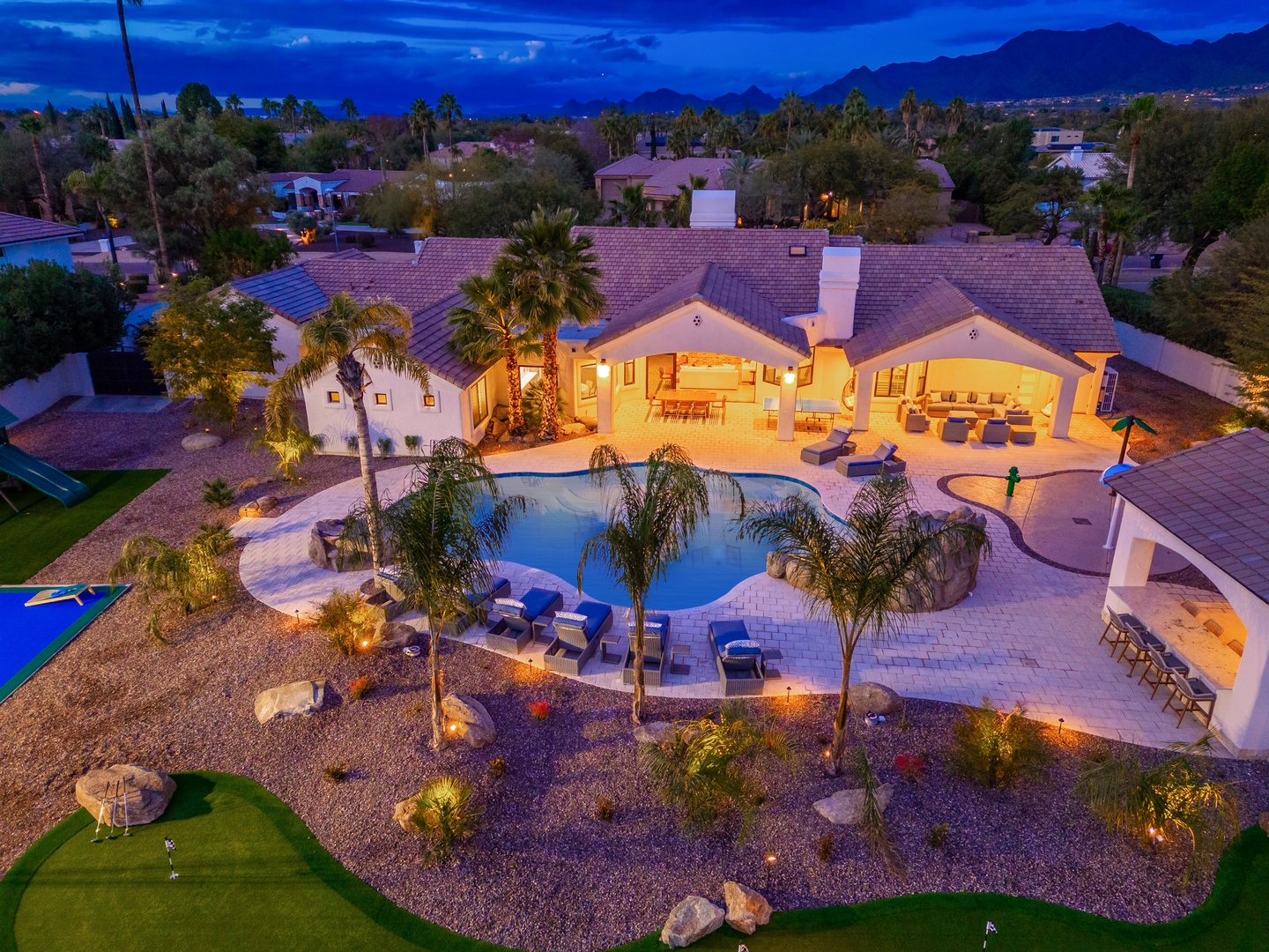 An aerial view captures the property at twilight with illuminated outdoor entertaining areas, a sparkling pool, and mountain backdrop in a palm-studded desert neighborhood.