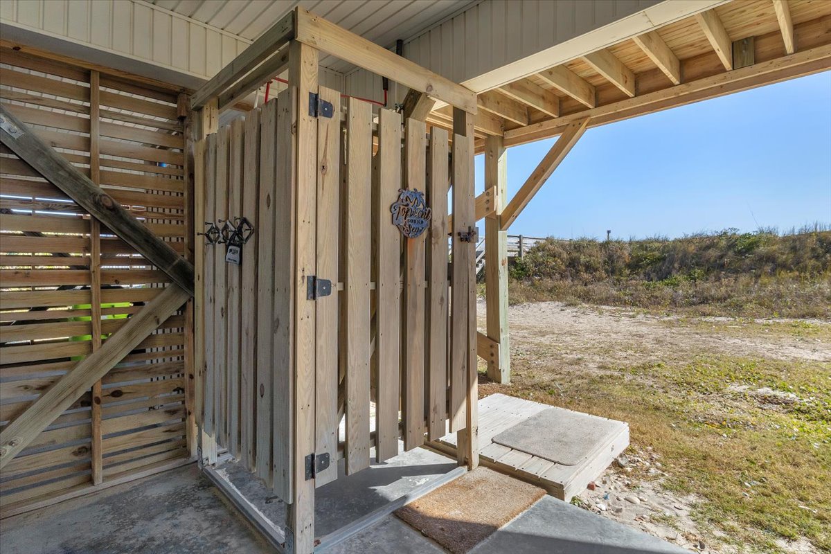 Rustic outdoor shower tucked beneath wooden stairs, offering privacy surrounded by coastal dunes and natural landscape.