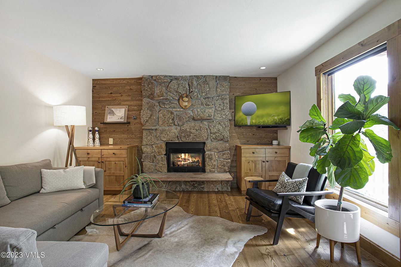 Relax in your mountain-inspired living room, where a stone fireplace creates a warm focal point and natural light pours through picture windows. Modern rustic furniture and lush greenery invite you to unwind in comfort.