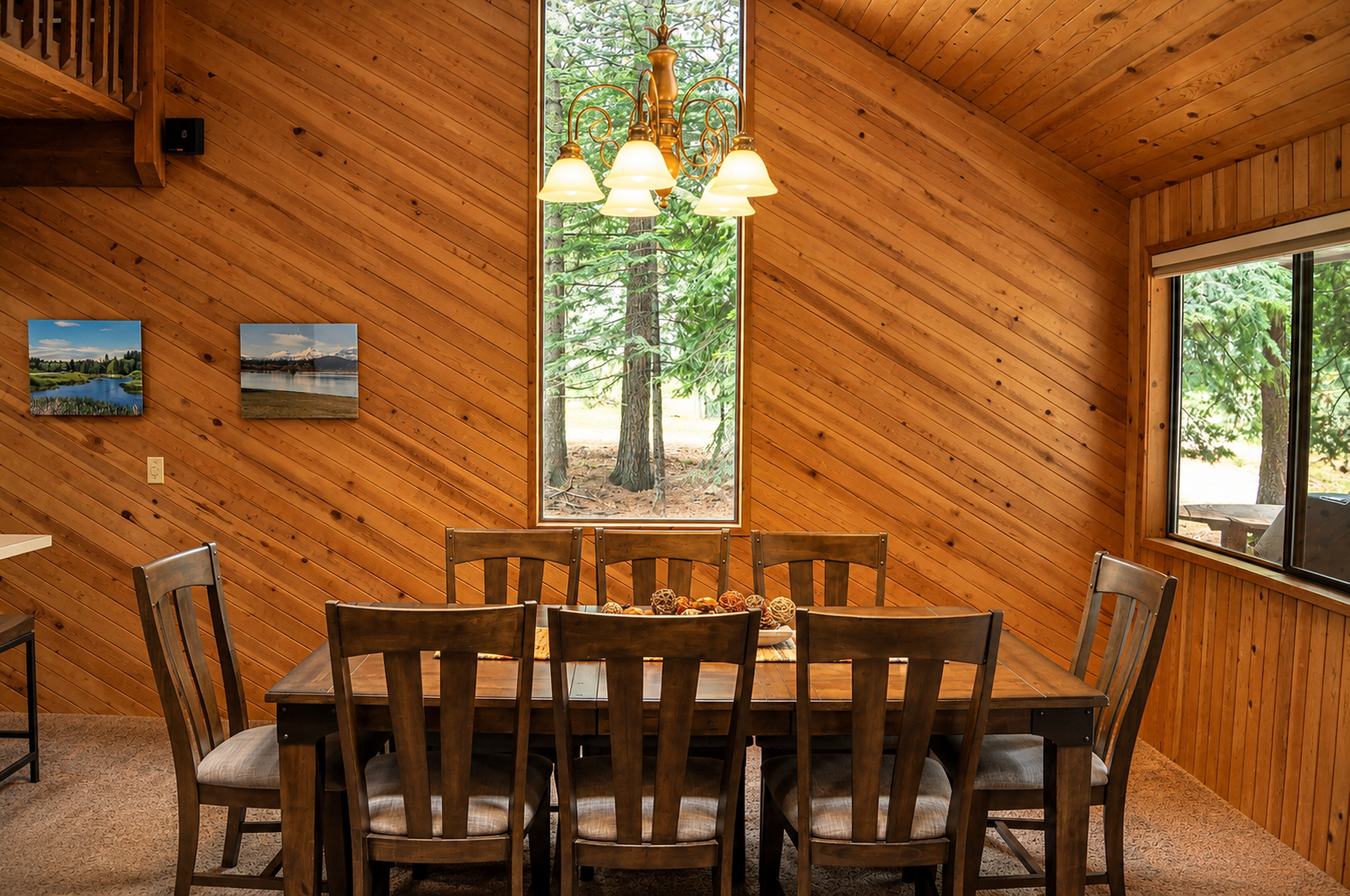 Gather around this inviting dining table surrounded by warm wood and forest views. The chandelier creates a cozy glow perfect for sharing meals and making memories together.