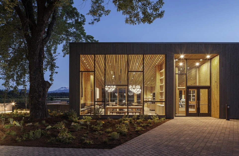 Modern architectural design showcases warm wood interiors through floor-to-ceiling windows, nestled among mature trees with mountain views.
