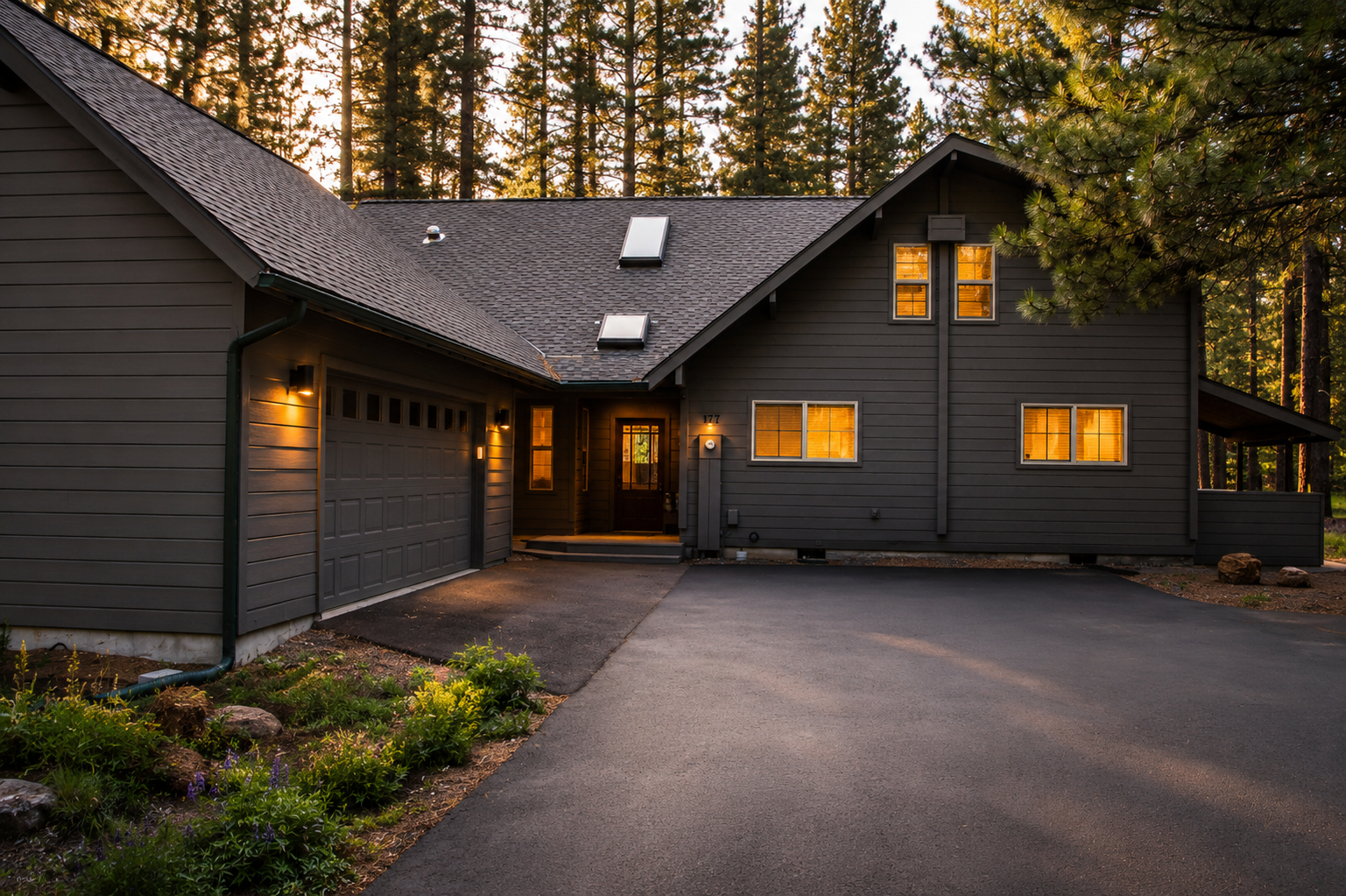 Contemporary mountain retreat nestled among towering pines, featuring a spacious driveway and welcoming entrance illuminated by warm evening light.