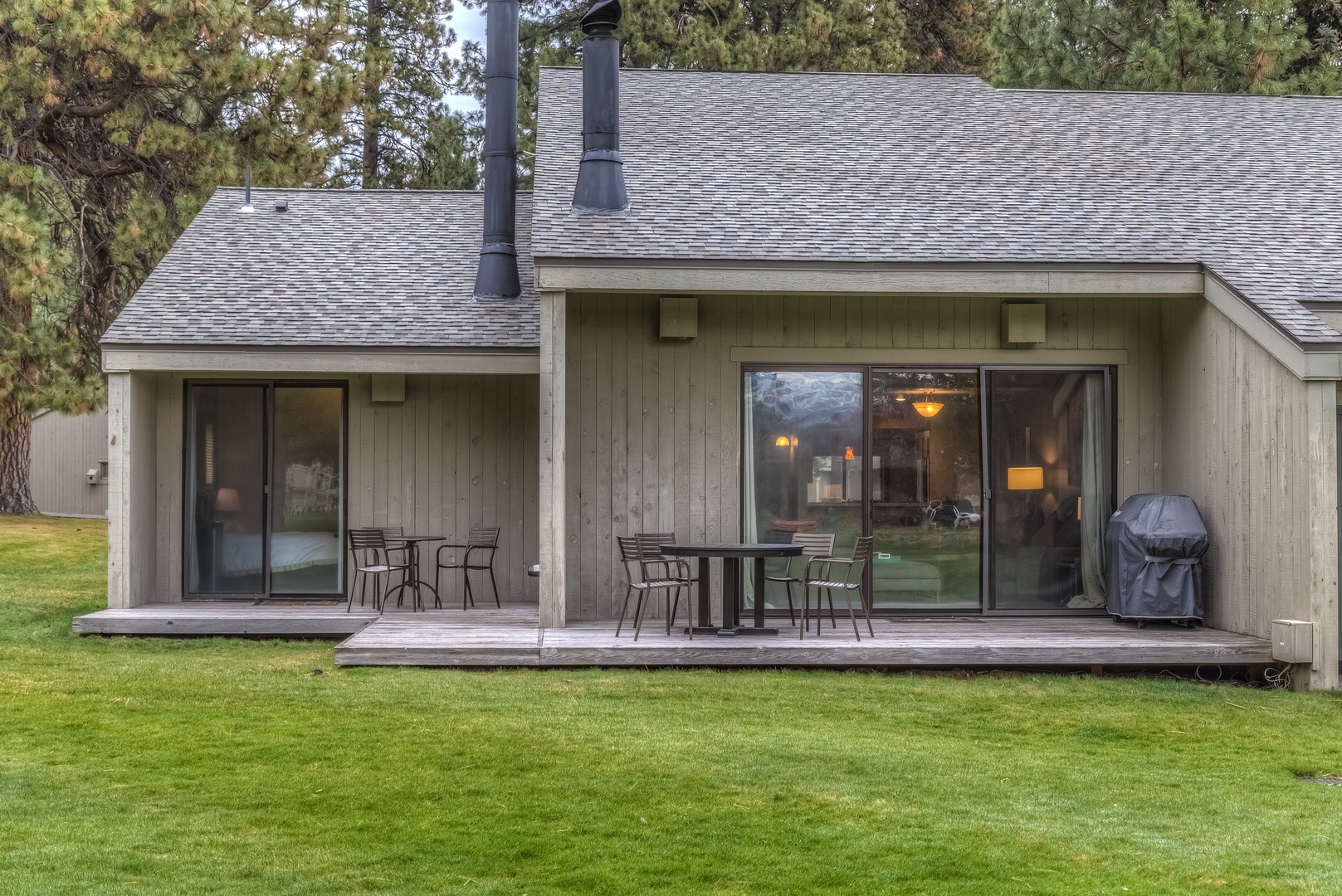 Modern vacation rental with spacious deck, outdoor dining, and BBQ setup surrounded by mature pine trees on manicured lawn.