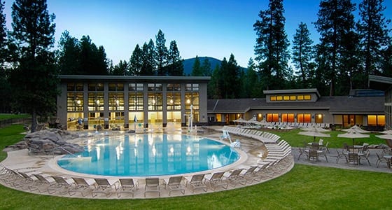 Modern resort facility with pristine circular pool surrounded by manicured lawns and towering evergreen trees.