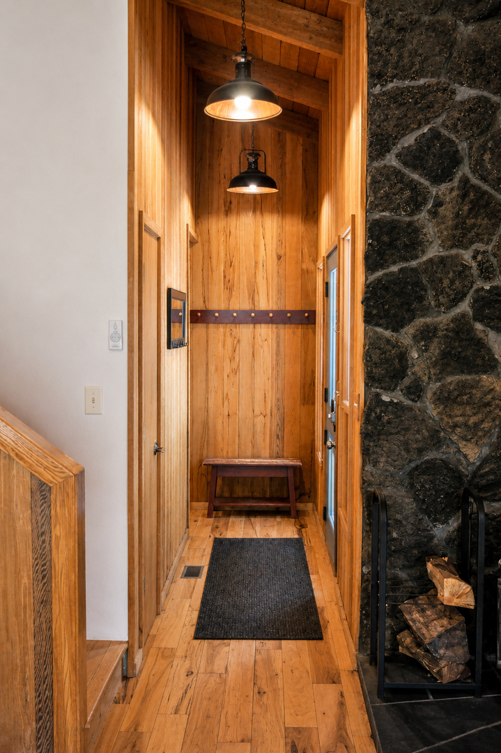 The property's inviting entryway features warm wood paneling, rustic pendant lighting, and a charming coat rack beside a stone fireplace stacked with firewood.