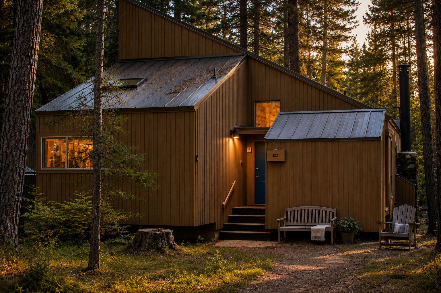 A modern cabin nestled among towering pines glows warmly as golden hour light filters through the forest, marking the peaceful entrance to this secluded woodland retreat.